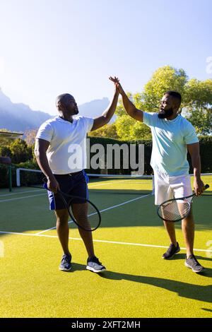 Tennis spielen, zwei verschiedene Freunde, die auf dem Platz im Freien hochfiebern, Schläger halten Stockfoto