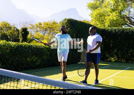 Tennis spielen, zwei verschiedene Freunde halten Schläger und unterhalten sich auf dem Platz im Freien Stockfoto