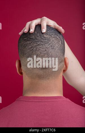 Die Frauenhand berührt sanft den Kopf eines Mannes von hinten und schafft ein Gefühl von Intimität und Verbindung auf einem leuchtend roten Hintergrund Stockfoto