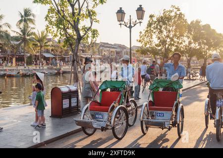 Hoi an (Hoian), Vietnam - 11. April 2018: Radfahrer-Rikschas (Trischas) und Touristen am Ufer des Flusses Thu Bon. Stockfoto