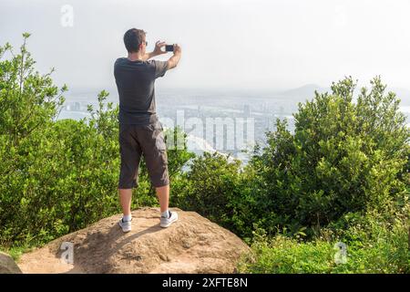 Mann mit Smartphone macht Fotos und genießt eine schöne Aussicht und frische Luft auf dem Berg zwischen grünen Büschen. Stockfoto