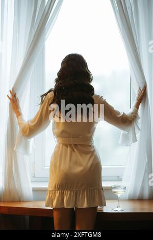 Die Braut macht sich morgens in ihrem Zimmer in einem Bademantel am Fenster fertig Stockfoto