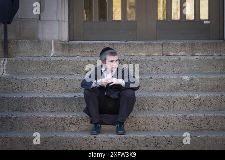 In der Pause sitzt ein junger orthodoxer jüdischer Junge auf seinen Schulstufen, verloren in seinen Gedanken. In Brooklyn, New York, März 2024. Stockfoto