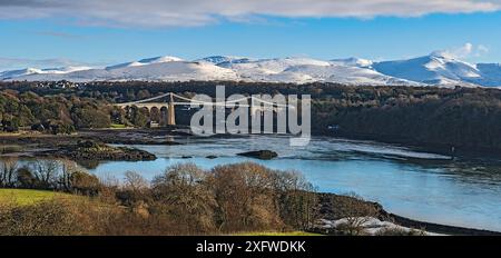 Menai Suspension Bridge, entworfen von Thomas Telford, von Anglesey aus gesehen über die Menai Strait, mit schneebedeckten Hügeln im Hintergrund. Nordwales, Großbritannien. Dezember 2017 Stockfoto