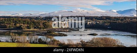 Menai Suspension Bridge, entworfen von Thomas Telford, von Anglesey aus gesehen über die Menai Strait, mit schneebedeckten Hügeln im Hintergrund. Nordwales, Großbritannien. Dezember 2017 Stockfoto