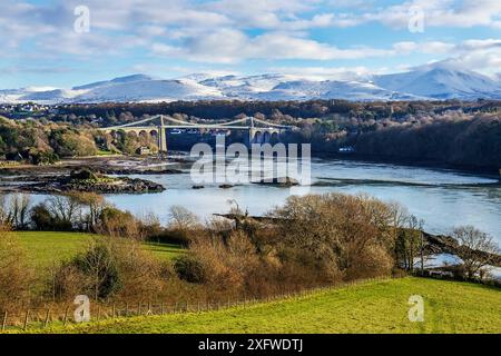 Menai Suspension Bridge, entworfen von Thomas Telford, von Anglesey aus gesehen über die Menai Strait, mit schneebedeckten Hügeln im Hintergrund. Nordwales, Großbritannien. Dezember 2017. Stockfoto