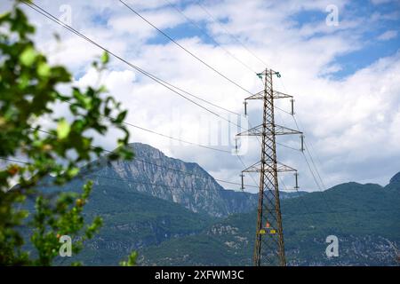 Arco, Trentino, Italien - 30. Juni 2024: Strompylon in Italien vor einer Berglandschaft und grünen Blättern von einem Baum. Symbolisches Bild von grünem Strom und nachhaltiger Energieversorgung *** Strommast in Italien vor einer Berglandschaft und grünen Blättern von einem Baum. Symbolbild grüner Strom und nachhaltige Energieversorgung Stockfoto