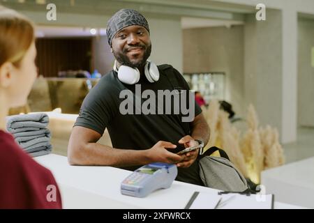 Junger afroamerikanischer Gym-Anwohner, der sich mit einer weiblichen Rezeptionistin in der riesigen Lobby des Fitnessclubs unterhält Stockfoto