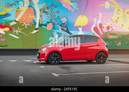 Rote Citroen C1 vor der farbenfrohen Wand. Seitenansicht des kompakten roten französischen Fließhecks in urbaner Umgebung - geparkt in der Nähe der Mauer mit volkstümlicher Kunst. Stockfoto