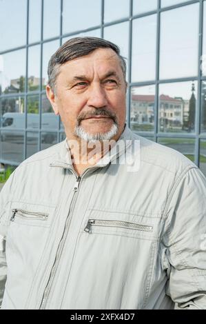 Ein attraktiver, jugendlicher, grauhaariger Mann von 70 Jahren vor dem Hintergrund von Bürogebäuden aus Spiegelglas. Porträt einer älteren grauhaarigen M Stockfoto