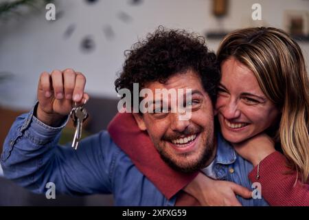 Fröhliches weißes junges Paar, das die Schlüssel ihres neuen Heims zeigt, umhüllt und lächelt. Stockfoto