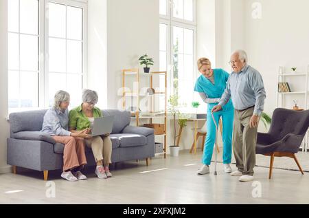 Die Krankenschwester im Altersheim hilft älteren Männern, mit dem Stock zu gehen, während zwei Frauen auf dem Sofa mit Laptop sitzen Stockfoto