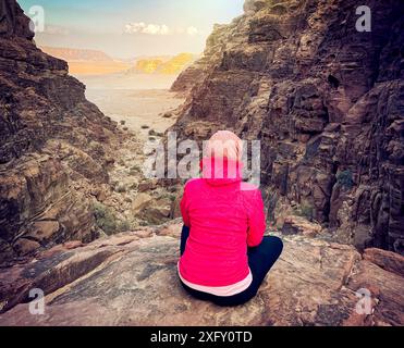Eine touristische Frau sitzt auf einem Steinvorsprung vor der Kulisse der Berge in der Wadi Rum Wüste bei Sonnenuntergang. Stockfoto