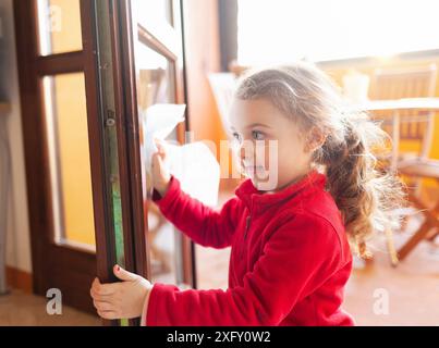 Das dreijährige Mädchen hilft bei der Haushaltsreinigung. Sie reinigt die Fenster mit einem Blatt Papier. Idee, die Zeit während der Kovid-19-Coronavirus-Quarantänezeit zu verstreichen. Stockfoto