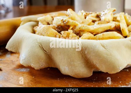 Torte machen. Gerollter Teig über eine Kuchenschale gelegt und mit Apfelscheiben gefüllt. Seitenansicht. Mehlkanister und Rollstift im Hintergrund. Stockfoto