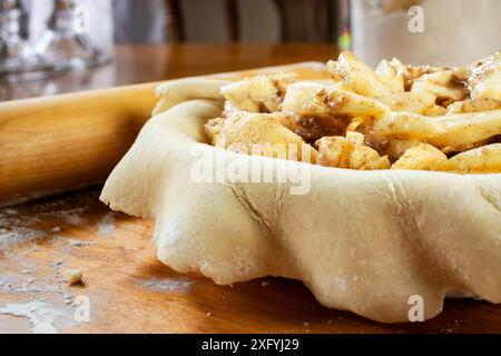 Torte machen. Gerollter Teig über eine Kuchenschale gelegt und mit Apfelscheiben gefüllt. Seitenansicht. Mehlkanister und Rollstift im Hintergrund. Stockfoto