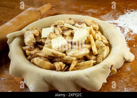 Torte machen. Gerollter Teig über eine Kuchenschale gelegt und mit Apfelscheiben gefüllt, die mit Butter gefüllt sind. Ansicht von oben. Rollstift im Hintergrund. Stockfoto