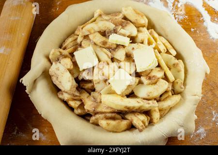 Torte machen. Gerollter Teig über eine Kuchenschale gelegt und mit Apfelscheiben gefüllt, die mit Butter gefüllt sind. Ansicht von oben nach unten. Rollstift im Hintergrund. Stockfoto