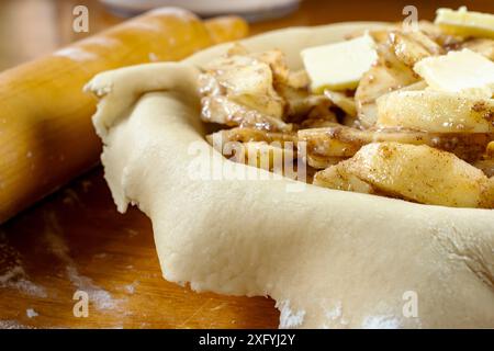 Torte machen. Gerollter Teig über eine Kuchenschale gelegt und mit Apfelscheiben gefüllt, die mit Butter gefüllt sind. Ansicht von oben. Rollstift im Hintergrund. Stockfoto