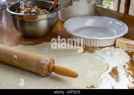 Torte machen. Teig auf einer bemehlten Holzoberfläche. Seitenansicht mit Nudelnadel, zubereiteten geschnittenen Äpfeln, weißer Tortenplatte, Butter und einem hölzernen rolli Stockfoto