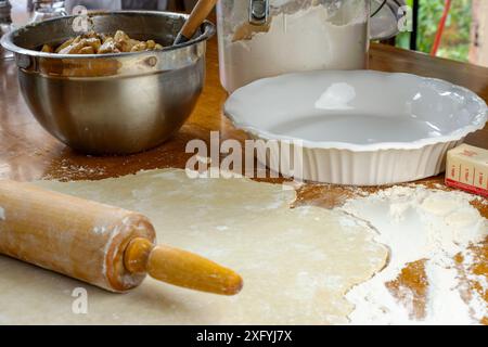 Torte machen. Teig auf einer bemehlten Holzoberfläche. Seitenansicht mit Nudelnadel, zubereiteten geschnittenen Äpfeln, weißer Tortenplatte, Butter und einem hölzernen rolli Stockfoto