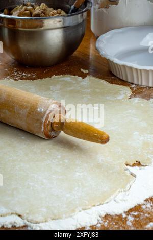 Torte machen. Teig auf einer bemehlten Holzoberfläche. Seitenansicht mit Nudelnadel, zubereiteten geschnittenen Äpfeln, weißer Tortenplatte, Mehlkanister und Woo Stockfoto