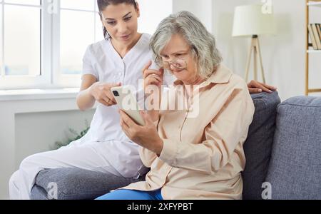 Junge fröhliche Krankenschwester zeigt älteren Frauen, wie man Smartphone benutzt. Stockfoto