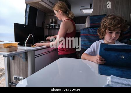 Eine Mutter kocht im Wohnmobil, während der Sohn mit einem digitalen Tablet spielt Stockfoto