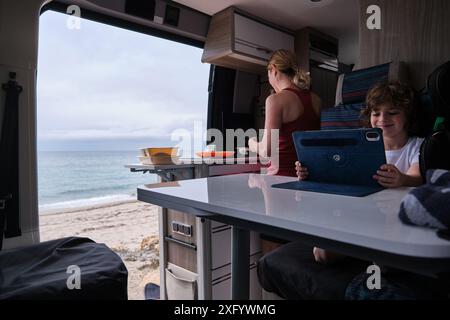 Eine Mutter kocht im Wohnmobil, während der Sohn mit einem digitalen Tablet spielt Stockfoto