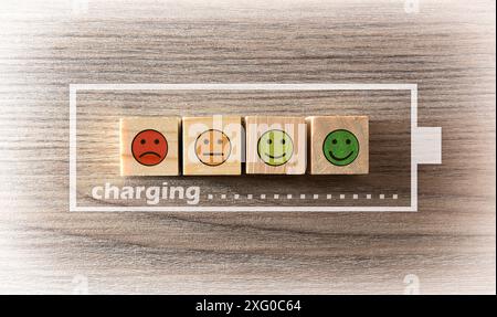 Konzept der Aufladung der Vitalenergie mit Holzblöcken mit Emoticons mit Ausdrücken auf Holztisch und Batteriesymbolen und Ladesymbolen. Stockfoto