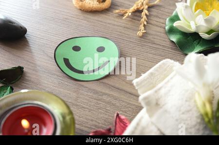 Positivität in der psychischen und körpermedizinischen Versorgung mit grünem Ausschnitt des lächelnden Gesichts auf Holztisch mit Elementen der Ruhe und Entspannung. Stockfoto