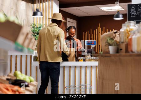 An der Kasse legt die afroamerikanische Verkäuferin Artikel in eine braune Papiertüte für den männlichen Kunden. Schwarzer Mann mit Hut, der an der Kasse wartet, während die Frau seine Produkte verpackt. Stockfoto