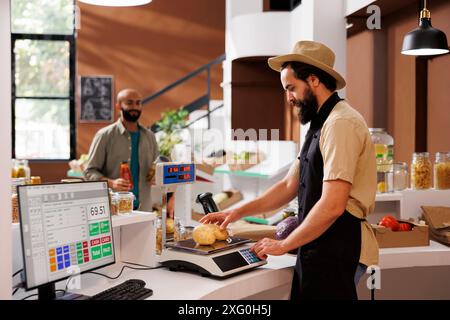 Kaukasischer Lagerhalter mit Hut und Schürze misst zwei Kartoffeln auf einer Waage. Der Kunde aus dem Nahen Osten geht in Richtung Theke, während der Anbieter frische Produkte aus der Region abwägt. Stockfoto