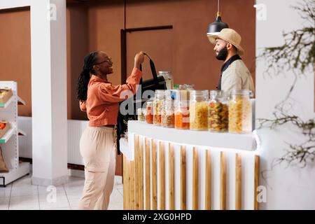 afroamerikanische Lieferfrau bereitet sich darauf vor, ein umweltfreundliches Geschäft zu verlassen, um frische, nachhaltige Artikel an Kunden zu liefern. Verkäufer und Kurierin interagieren am Kassenschalter. Stockfoto