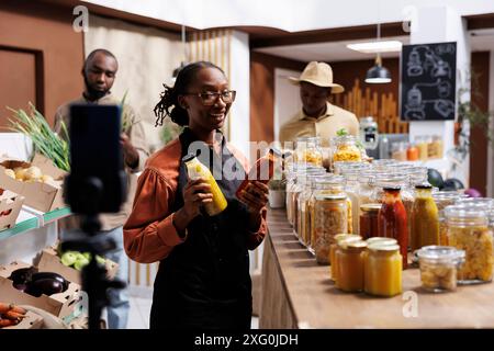 Schwarze weibliche Besitzerin wirbt für umweltfreundliche Produkte in einem Zero Waste Shop vor Ort. Afroamerikanische Frau mit Gläsern filmt ein Video mit einem Smartphone, in dem frische, biologische Lebensmittel in Regalen gezeigt werden. Stockfoto