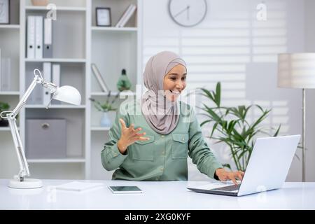Glückliche Frau, die einen Hijab trägt und einen Online-Videoanruf in einer modernen Home-Office-Umgebung führt. Sie lächelt und gibt Gesten, während sie einen Laptop auf einem weißen Schreibtisch mit Tablet und Lampe benutzt. Stockfoto