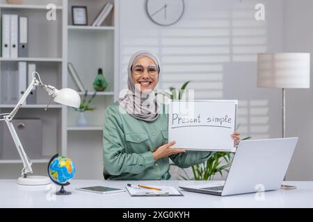 Lehrerin mit Hijab und Brille, die einen Online-Kurs über die gegenwärtige Simple-Form führt. Mit Laptop, Whiteboard und Lehrmaterial am Schreibtisch eines Heimbüros. Stockfoto