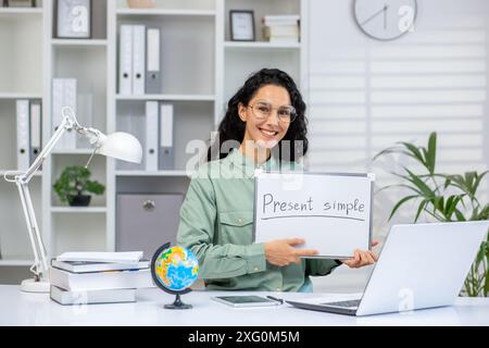 Ein lächelnder Lehrer hält ein Whiteboard mit der Aufschrift „Präsent Simple“. Die Szene spielt in einem modernen Büro mit einem Laptop, Büchern und einem Globus, was auf einen Online-Englischkurs hinweist. Stockfoto