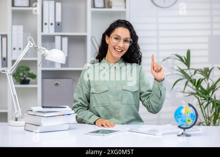 Fröhliche Lehrerin hebt ihre Hand, während sie an einem Schreibtisch in einem modernen, organisierten Klassenzimmer sitzt. Umgeben von Büchern, einem Globus und Bürobedarf strahlt sie Begeisterung und Wissen aus. Stockfoto