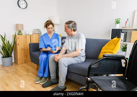 Junge Ärztin in blauer Uniform besucht einen behinderten Seniorenmann zu Hause Stockfoto