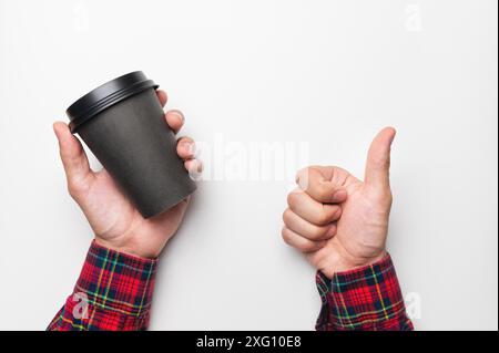 Die Hand eines Mannes hält eine leere papierschwarze Tasse Kaffee auf weißem Hintergrund in der einen Hand und zeigt mit der anderen Hand eine Geste der Klasse, Daumen hoch Stockfoto