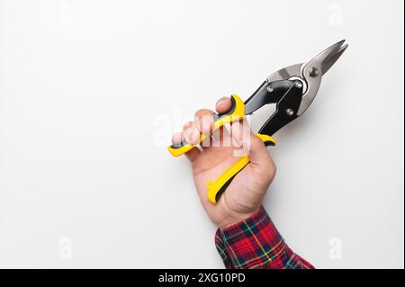 Die Hand eines Mannes in einem karierten Hemd hält ein Bauwerkzeug auf weißem Hintergrund, eine hellgelbe Schere zum Schneiden von Metall, Nahaufnahme von oben Stockfoto