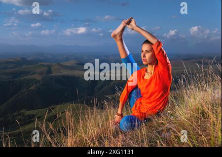 Junge attraktive Yogi-Frau, die Yoga praktiziert, Variationen von Krounchasana-Übungen macht, Reiher-Pose macht, Sport in der Natur macht, Sportbekleidung trägt Stockfoto