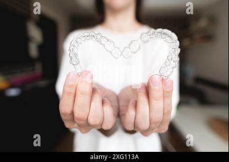 Zahnärztliche Versorgung. Nahaufnahme transparenter Aligner in den Händen eines Mädchens, das steht und ein herzförmiges kieferorthopädisches Gerät vor der Kamera zeigt. Stockfoto