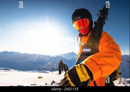 Foto eines Athleten in Helm und Maske mit Skiern auf der Schulter. Ein junger Mann steht in den Bergen vor dem Hintergrund der Sonne und schaut zu Stockfoto