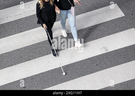 Die Szene einer blinden Frau, die auf einer Zebrakreuzung läuft, wurde von einer anderen Person mit ihrem weißen Stock unterstützt. Hilfe in den frühen Stadien der Blindheit Stockfoto
