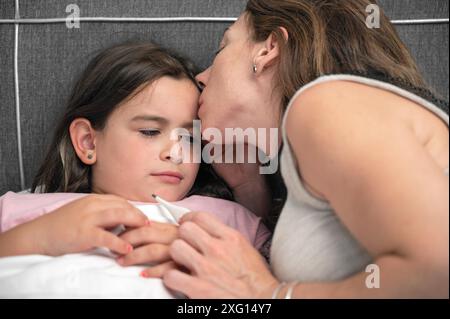 Mutter misst die Temperatur ihrer kranken Tochter. Krankes Kind mit hohem Fieber liegt im Bett und Mutter hält Thermometer. Hochwertige Fotos Stockfoto