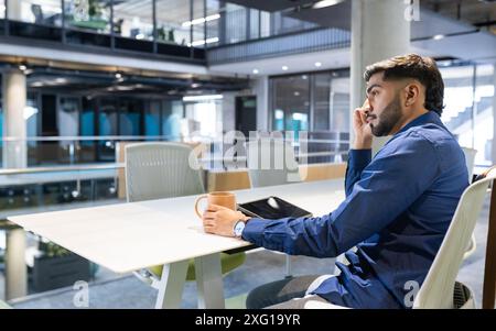 Sprechen auf dem Smartphone, Mann hält eine Kaffeetasse und sitzt im modernen Büro, Kopierraum Stockfoto