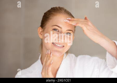 Hautpflege anwenden, Frau in weißem Gewand lächelt und ihr Gesicht berührt Stockfoto