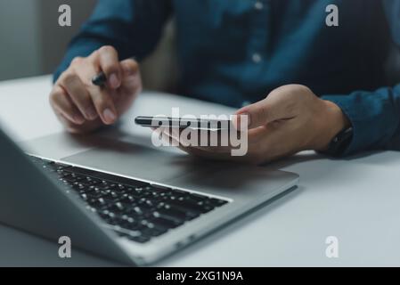 Ein Mann benutzt einen Laptop und ein Handy. Er hält das Handy in der Hand und schaut auf den Laptop Stockfoto